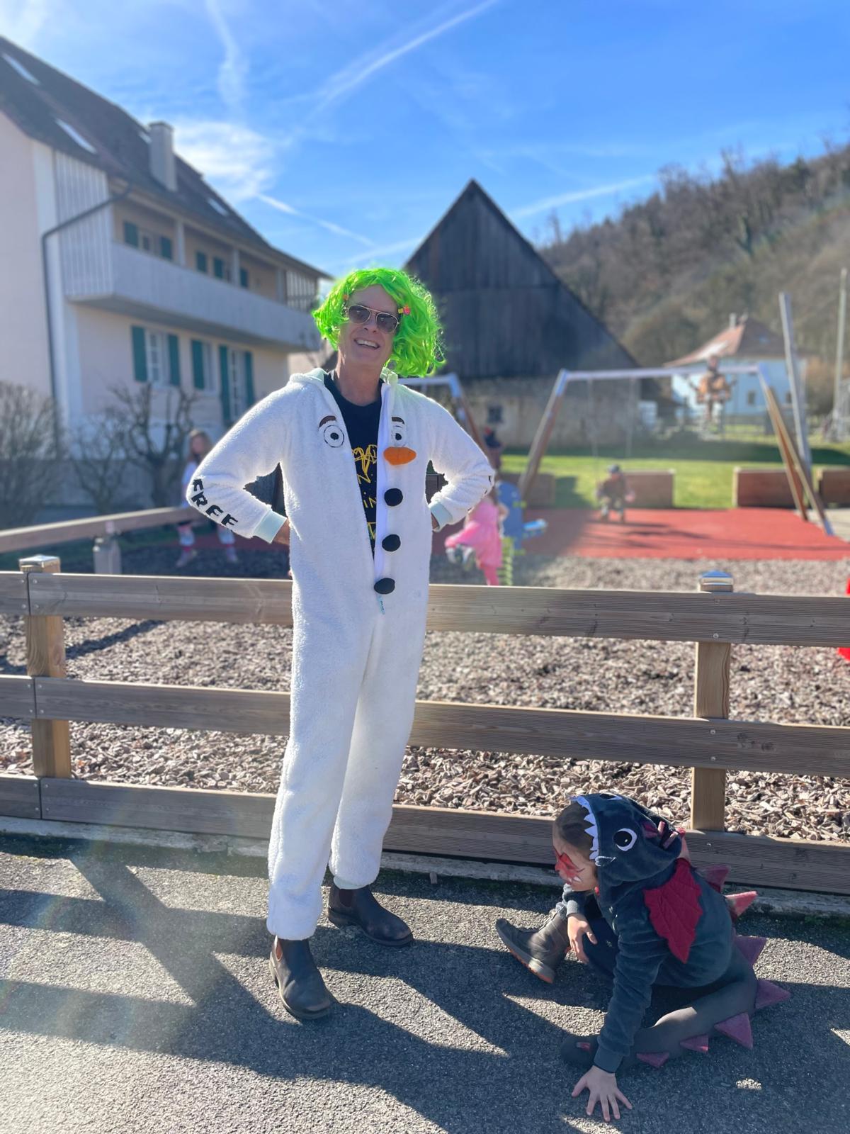 Jeremy in a snowman onesie and green wig with a child in a dinosaur costume